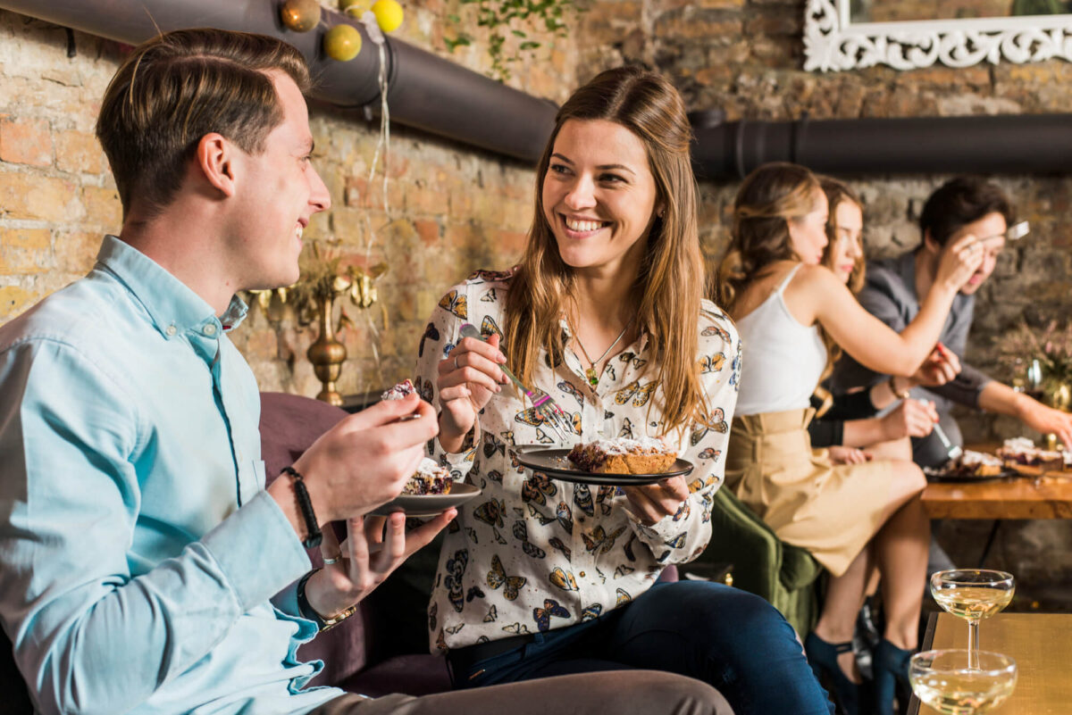 Um restaurante cheio. Na imagem, um homem e uma mulher conversam, em uma mesa mais ao fundo é possível visualizar outro grupo de pessoas conversando. Um ótimo local para painéis que melhoram a acústica.