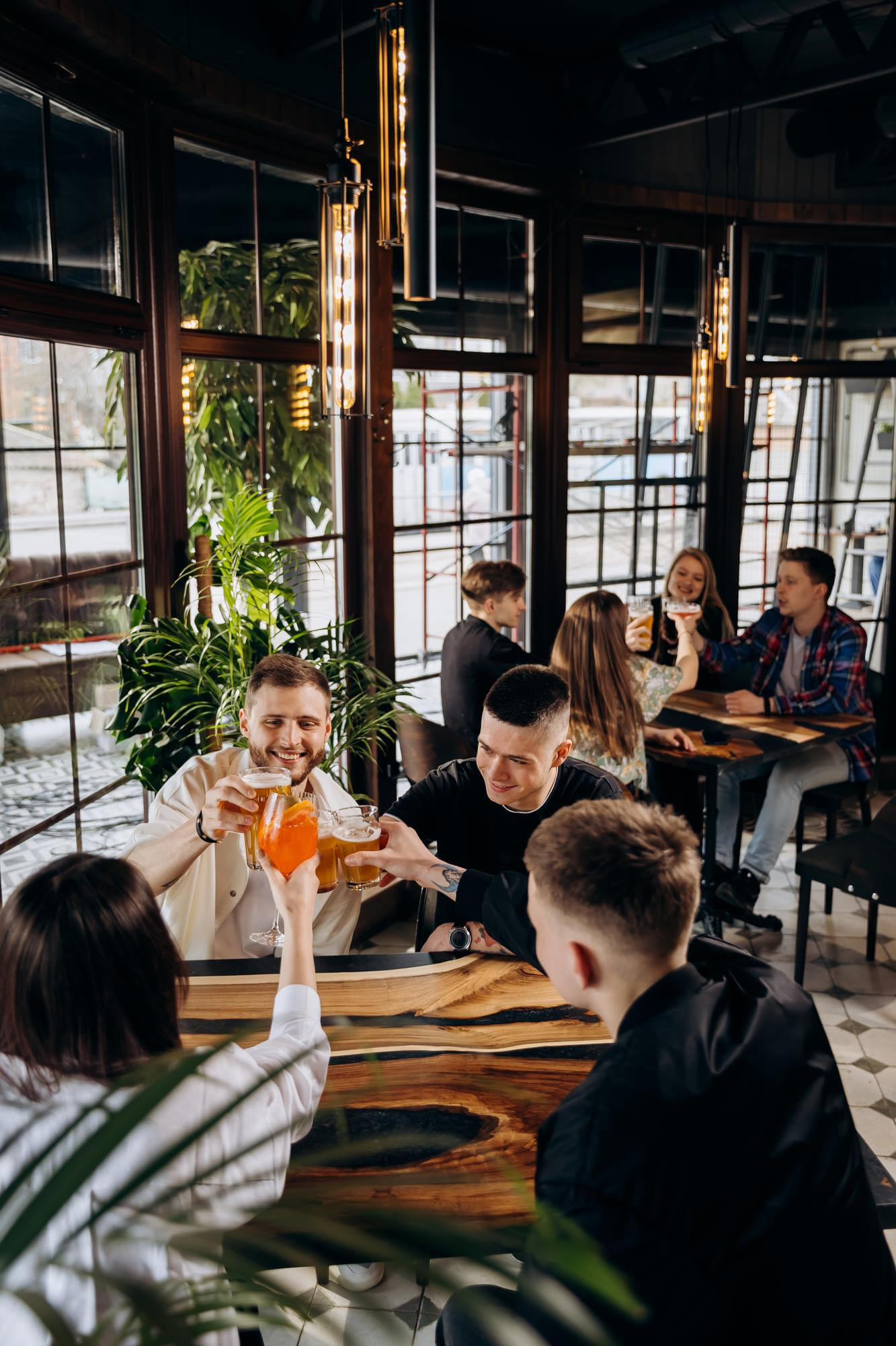 Um restaurante moderno, com tons amadeirados e escuros. Na mesa, pessoas brindam, com drinks. Um ótimo local para painéis que melhoram a acústica.